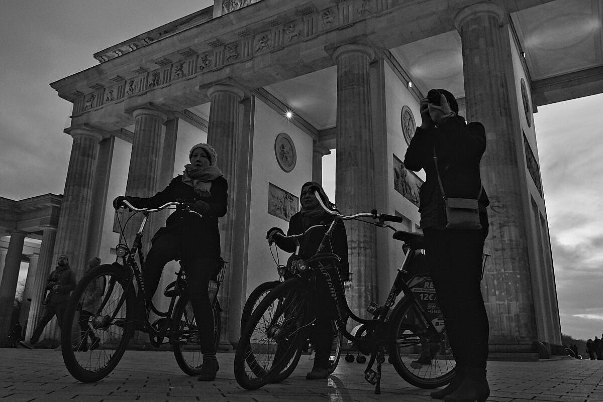 Three women on bicycles at the Brandenburger Tor in Berlin, around sunset.